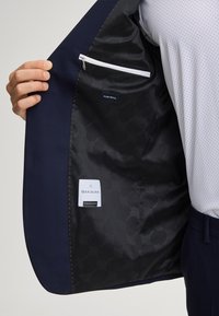 Blazer in lana blu navy con fodera nera a motivo, dotato di tasca con cerniera e etichette cucite con informazioni sul tessuto e marchio, che mostrano un design strutturato.