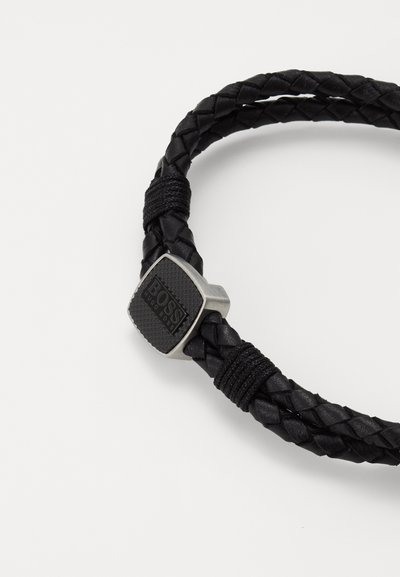 Bracelet en cuir tressé noir avec un fermoir en métal argenté et une plaque logo texturée. Les détails incluent des enroulements en cordon noir.