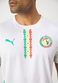 Bela športna majica z zelenim logotipom Puma, barvitimi vertikalnimi vzorci v zeleni, rumeni in rdeči ter vezenim grbom z levom.