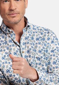 Chemise à manches longues, blanche avec un motif floral bleu, vert et orange. Présente un col et des boutons sur le devant.