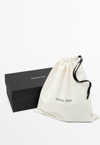 Bílý bavlněný sáček na stažení s nápisem "Massimo Dutti" vytištěným černě, vedle černé obdélníkové krabičky. Jednoduchý design, hladká textura.
