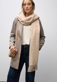 Beige uldhalstørklæde draperet over en lys grå frakke med blød tekstur. Kombineret med mørke jeans og en lille beige crossbody taske.
