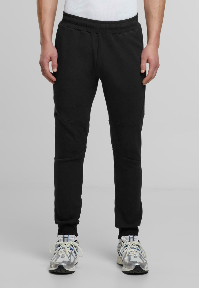 Svarta joggingsbyxor med elastisk midja och avsmalnande ben. Har sömdetaljer vid knäna. Kombineras med grå och vita löparskor.
