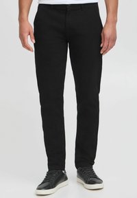 Schwarze Baumwollhosen mit schlanker Passform, ausgestattet mit Gürtelschlaufen, Vordertaschen und einem sauberen Finish. Tragen mit schwarzen Sneakers.