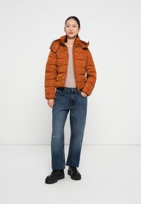 Brun quiltet pufferjakke med hætte, parret med lysebeige sweater og blå vide jeans. Sorte chunky sko. Neutral baggrund.