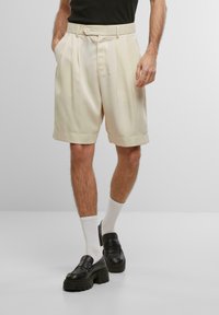 Shorts plissés de couleur crème avec des poches latérales, une fermeture à bouton et des ourlets repliés, associés à des mocassins à plateforme noirs et des chaussettes blanches.