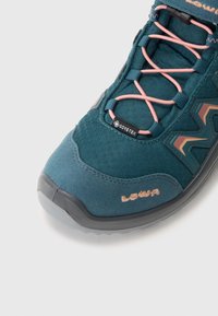 Türkiz túracipő rózsaszín fűzőkkel, GORE-TEX címkével, mintás oldal díszítéssel, gumírozott orral és texturált felső anyaggal. Tartós kialakítás szabadtéri használatra.