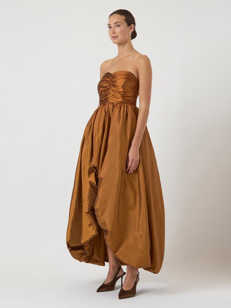 Woman wearing a strapless, ruched, copper-colored voluminous midi dress paired with pointed-toe high-heeled slingback shoes, standing on white background.