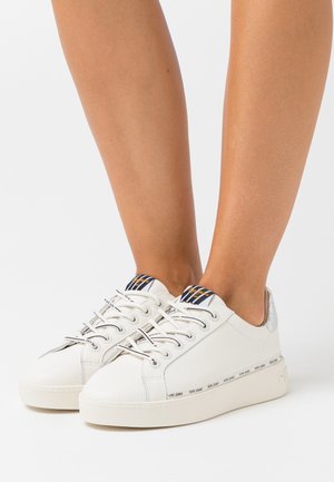 Baskets basses blanches avec langue rayée noir et blanc, détail argenté au talon et lacets à la marque, portées pieds nus.