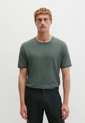 Jeune homme aux cheveux blonds courts, portant un t-shirt vert col rond et un pantalon noir, debout devant un fond blanc uni.