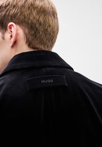 Μαύρο βελούδινο παλτό με καπιτονέ γιακά, ορατή ραφή και ορθογώνια ετικέτα κεντημένη με το "HUGO" στην πλάτη.