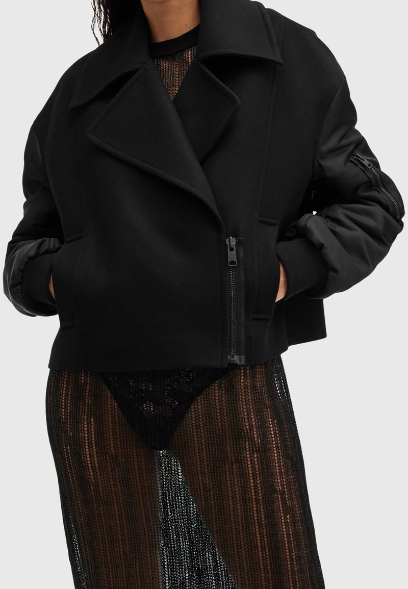 Veste noire oversize avec fermeture éclair et poches portée sur une robe en dentelle noire transparente, mains glissées dans les poches de la veste, fond gris.