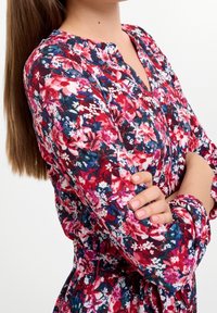 Vestido floral con un patrón rojo y azul, que presenta un escote abotonado, mangas largas y detalles fruncidos en la cintura. Tela texturizada.