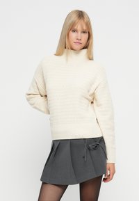 Pull en tricot crème avec un col haut, motif texturé et silhouette ample, associé à une jupe plissée gris foncé et des collants noirs.