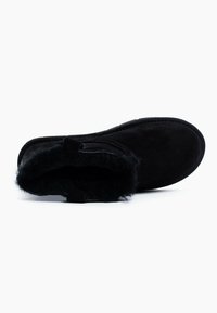 Botín de ante negro con un suave forro de piel. Presenta una punta redondeada, una pequeña asa y una suela plana. Textura suave y diseño minimalista.