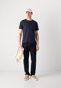 Marineblå t-skjorte med en subtil logo, marineblå bukser, beige sneakers og en lyse farget genser lagt over den ene armen. Nøytral bakgrunn.