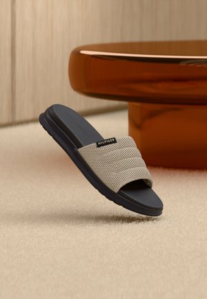 Beige Mesh-Sandale zum Hineinschlüpfen mit schwarzer Sohle und Hilfiger-Label, auf beigem Teppich nahe einem runden, bernsteinfarbenen Tischfuß platziert.