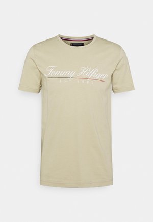 Beiges kurzärmeliges Baumwoll-T-Shirt mit "Tommy Hilfiger EST. 1985" in weißer Schrift über der Brust gedruckt.
