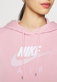 Ljusrosa Nike-hoodie med dragsko, som har en stor vit logotyp och broderade detaljer. Mjukt tyg med slät textur.