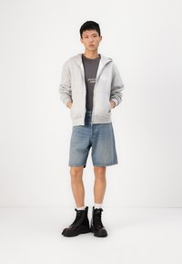 Sweat à capuche zippé gris, t-shirt graphique noir, short en denim bleu et bottes de combat noires avec des accents rouges. Tissu lisse et coupe décontractée.