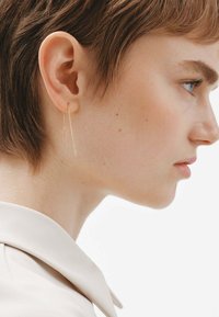 Pendientes colgantes en tono dorado con cadenas delgadas y alargadas. Su diseño sencillo complementa la forma de la oreja, colgando justo por debajo del lóbulo.
