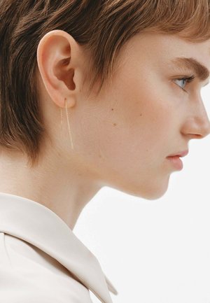 Pendientes colgantes en tono dorado con cadenas delgadas y alargadas. Su diseño sencillo complementa la forma de la oreja, colgando justo por debajo del lóbulo.