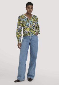 Una donna indossa una blusa a maniche lunghe floreale multicolore, jeans a vita alta e a gamba larga blu, e scarpe scure con punta. Sta in piedi con una mano nella tasca.