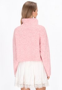 Gekürzter rosa Pullover in gerippter Textur mit hohem Kragen. Weiches Material und lockere Passform, getragen über einem weißen Kleid mit Spitzenbesatz.