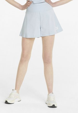 Person iført lyseblå træningsshorts og hvide sneakers, stående med hænderne på hofterne mod en hvid baggrund.