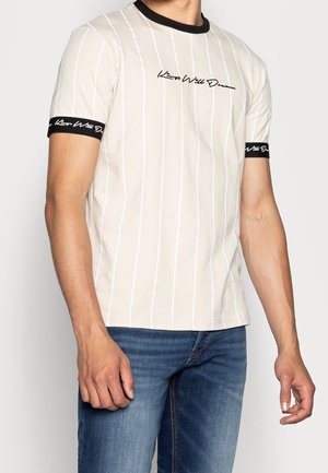 Uomo che indossa una T-shirt beige a righe verticali con colletto nero e fasce sulle maniche con testo in corsivo bianco, abbinata a un paio di jeans blu.