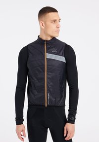 Sort quiltet vest med lynlås fortil og grå refleksstribe; har logo og teksturerede paneler. Bæres over en sort langærmet top.