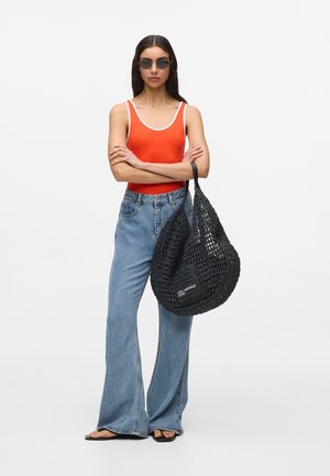 Žena v oranžovom tielku bez rukávov, modrých džínsach s širokými nohavicami, v čiernych slnečných okuliaroch, drží veľkú čiernu tkanú kabelku, stojí so založenými ramenami.