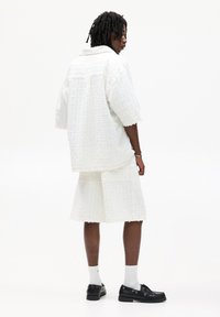 Camicia bianca a maniche corte con texture e pantaloni corti abbinati, realizzati in un tessuto leggero. Abbinata a scarpe nere e calzini bianchi.