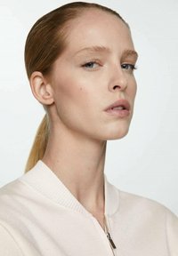 Model with light skin, straight hair tied back, wearing a cream-colored bomber jacket with a ribbed collar and zipper detail.