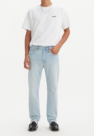 Homme portant un t-shirt blanc Levi's, un jean bleu clair, des mocassins en cuir noir et un collier chaîne argenté, debout avec une main dans la poche.