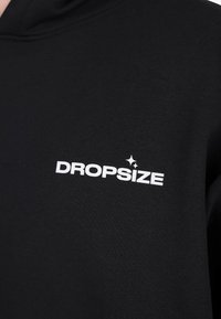 Tessuto nero con il testo bianco "DROPSIZE" e due piccole icone a forma di stella sopra le lettere "S" e "I" nella zona del petto.