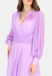 Liliowa sukienka z głębokim dekoltem w kształcie V i długimi, bufiastymi przezroczystymi rękawami. Posiada marszczoną talię oraz piórkowy akcent na ramieniu. Gładka faktura.