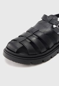 Crna kožna sandala s tkane trakama, debelim gaznim slojem, podesivom remenom oko gležnja i utisnutim Calvin Klein logotipom na gornjem dijelu.