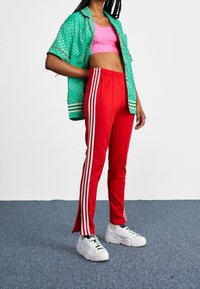 Grünes, gemustertes Kurzarmhemd, pinkes Crop-Top, rote Jogginghose mit weißen Streifen und weiße Turnschuhe mit strukturiertem Profil.
