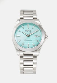 Montre en acier inoxydable avec un cadran rayé turquoise, des index d'heure argentés et un affichage de date. Elle comprend un bracelet métallique robuste et est résistante à l'eau.