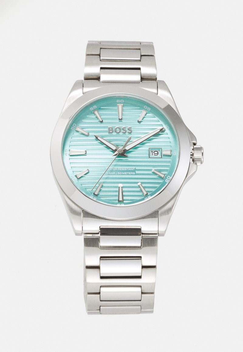 Montre en acier inoxydable avec un cadran rayé turquoise, des index d'heure argentés et un affichage de date. Elle comprend un bracelet métallique robuste et est résistante à l'eau.