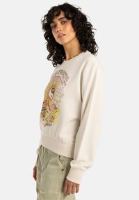 Beige sweatshirt med rund halsringning, ribbade ärmslut och en jordnära grafisk design med blommor, ett yin-yang-symbol och livfulla färger.