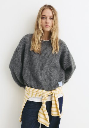 Maglione grigio lavorato a maglia con maniche larghe, indossato sopra una camicia bianca e jeans blu. Tessuto a righe gialle e bianche annodato in vita.