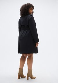 Tengerészkék ballonkabát öves derékrésszel, hátsó sliccel és széles gallérral, párosítva barnás színű bokacsizmával, melynek texturált felülete és blokk sarokja van.