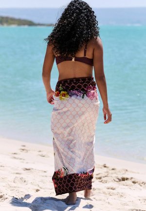 Kvinde med krøllet hår, der går barfodet på en sandstrand iført blomsterfarvet sarong og mørk bikini top, vendt mod det rolige blå hav.