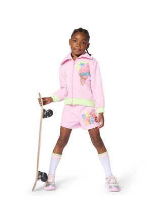 Jong meisje in roze korte broek en jas houdt een skateboard vast, staat met gespreide benen tegen een witte achtergrond.