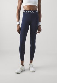 Granatowe legginsy Nike Pro z szerokim, markowym pasem, wykonane z elastycznego materiału. Noszone z białym krótkim topem i białymi butami sportowymi.