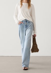 Top a maniche lunghe beige chiaro indossato sopra jeans larghi a vita alta di colore blu chiaro. La modella tiene una borsa in suede marrone con una tasca.