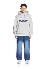 Szary hoodie z logo "PROSTO." oraz kieszenią z przodu, w zestawieniu z luźnymi niebieskimi dżinsami. Model nosi jasne sneakersy, na tle neutralnym.