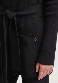 Close-up van een persoon die een zwarte gebreide cardigan draagt, geknoopt in de taille, met een voorzak en gouden ringen om hun hand.